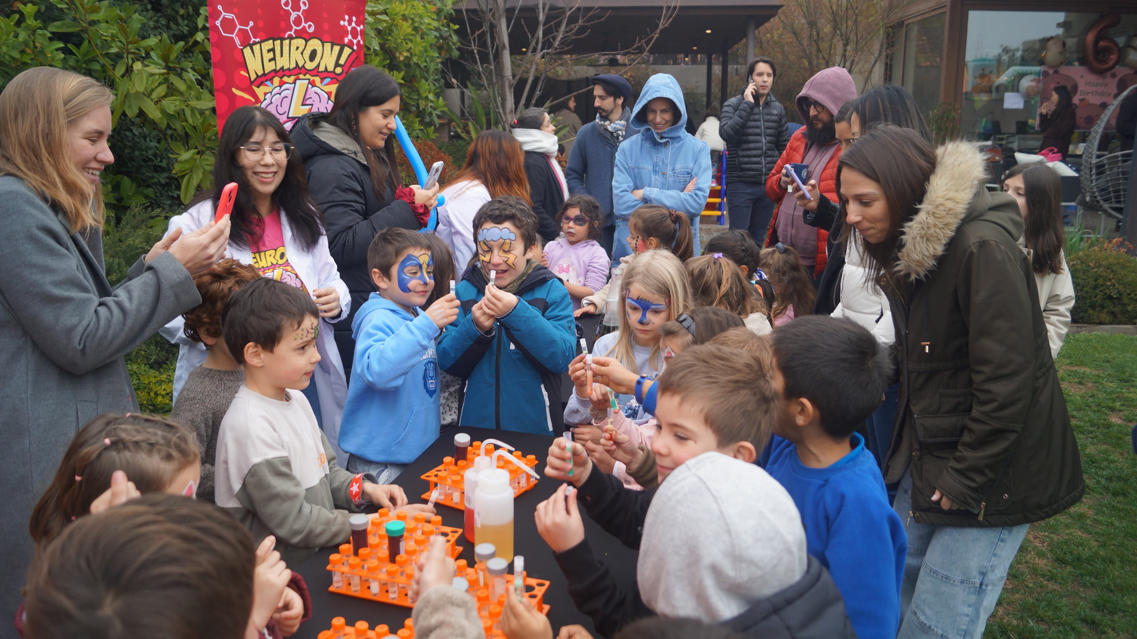 Niños con la cara pintada observan sus tubos de ensayo junto a educadoras con bata y un banner de Neuron