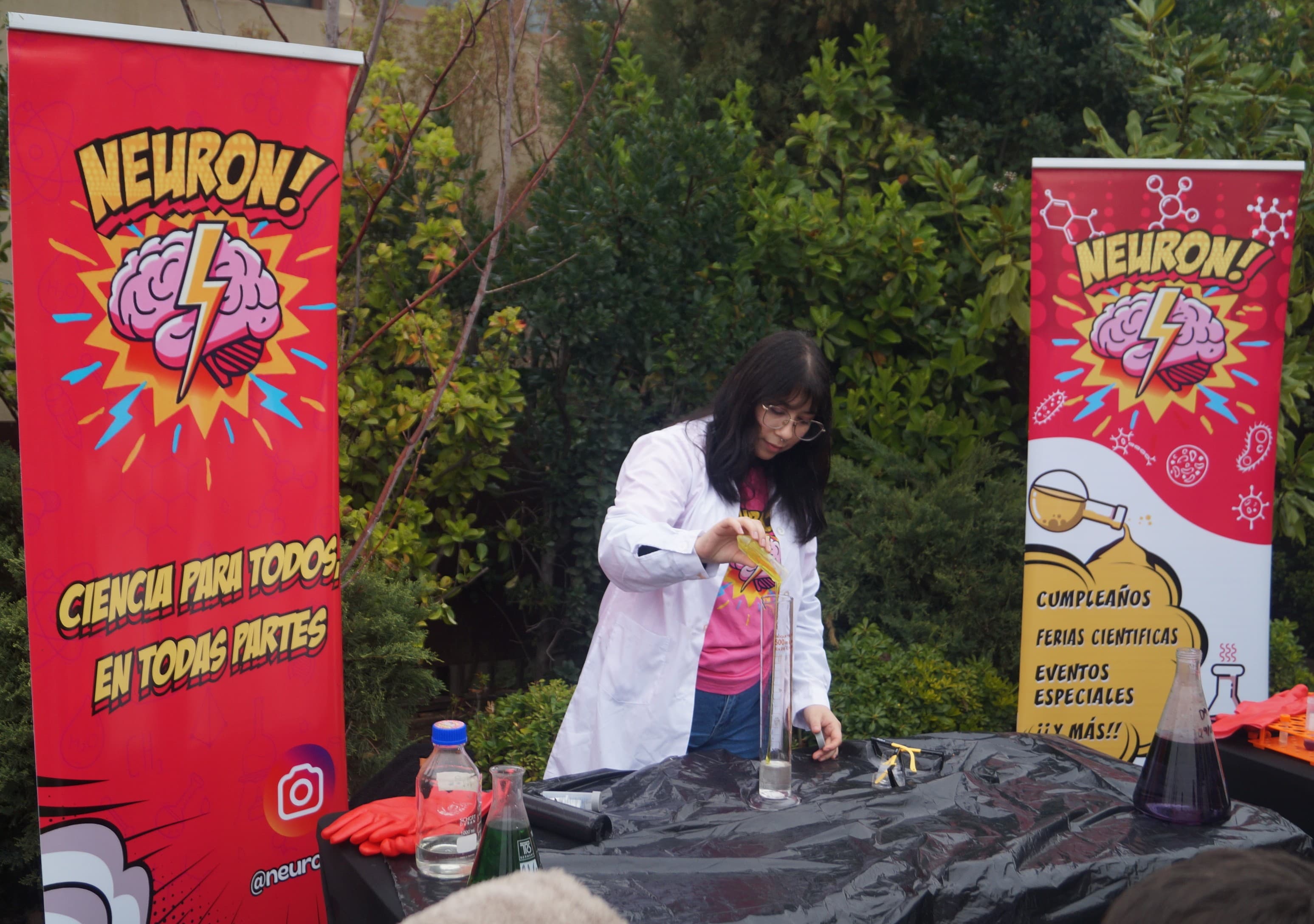 Educadora con bata vierte un líquido de colores en una probeta entre dos banners de Neuron